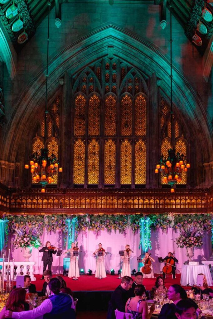 String Ensemble — Performance in a Gothic Great Hall Five Urban Soul Orchestra string players performing on a floral-decorated stage beneath a large Gothic window.