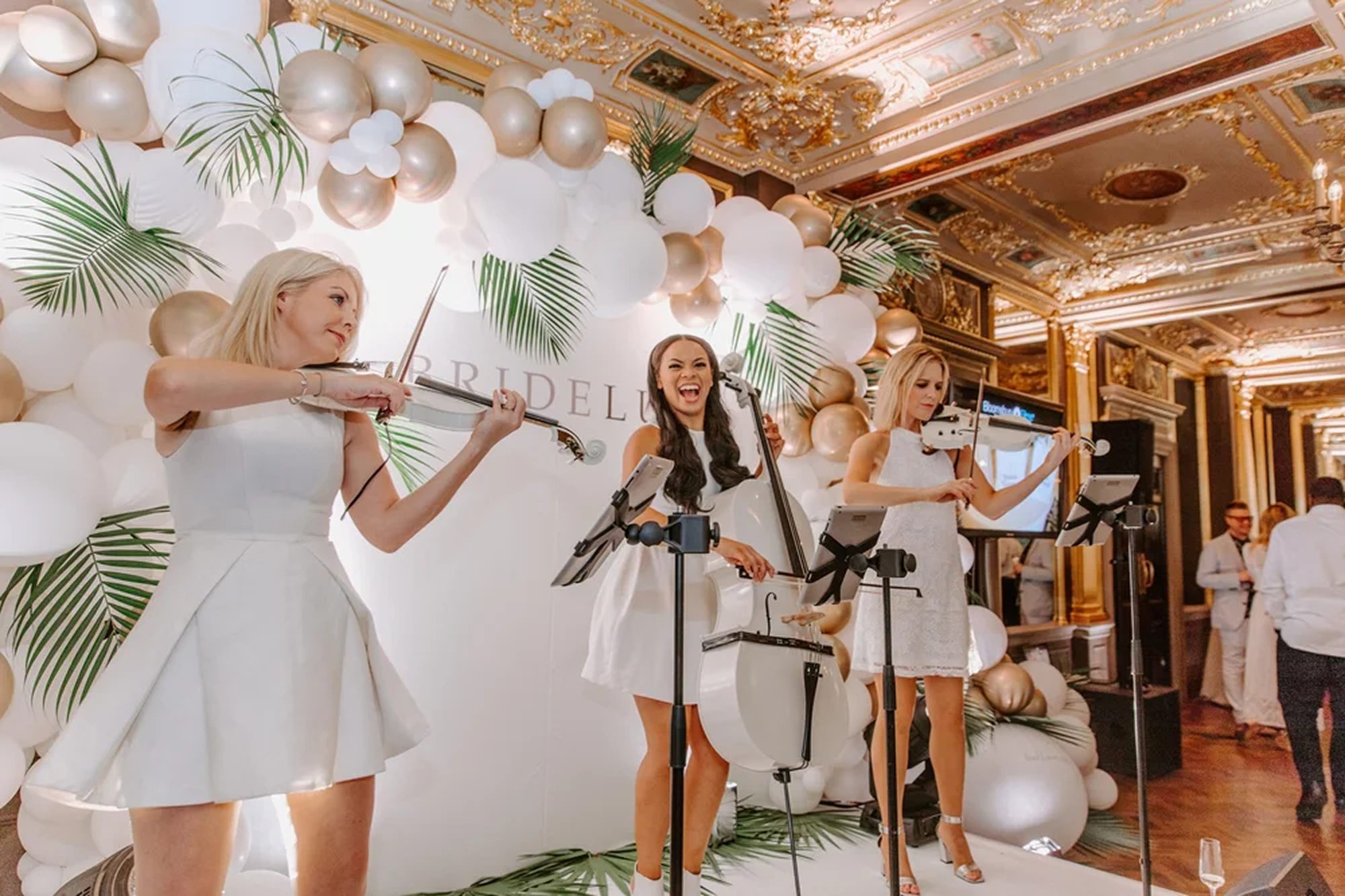 Electric Strings Performing at a Luxury Venue Event Three Urban Soul Orchestra string players in white performing on electric violins and cello against a white balloon and palm installation.
