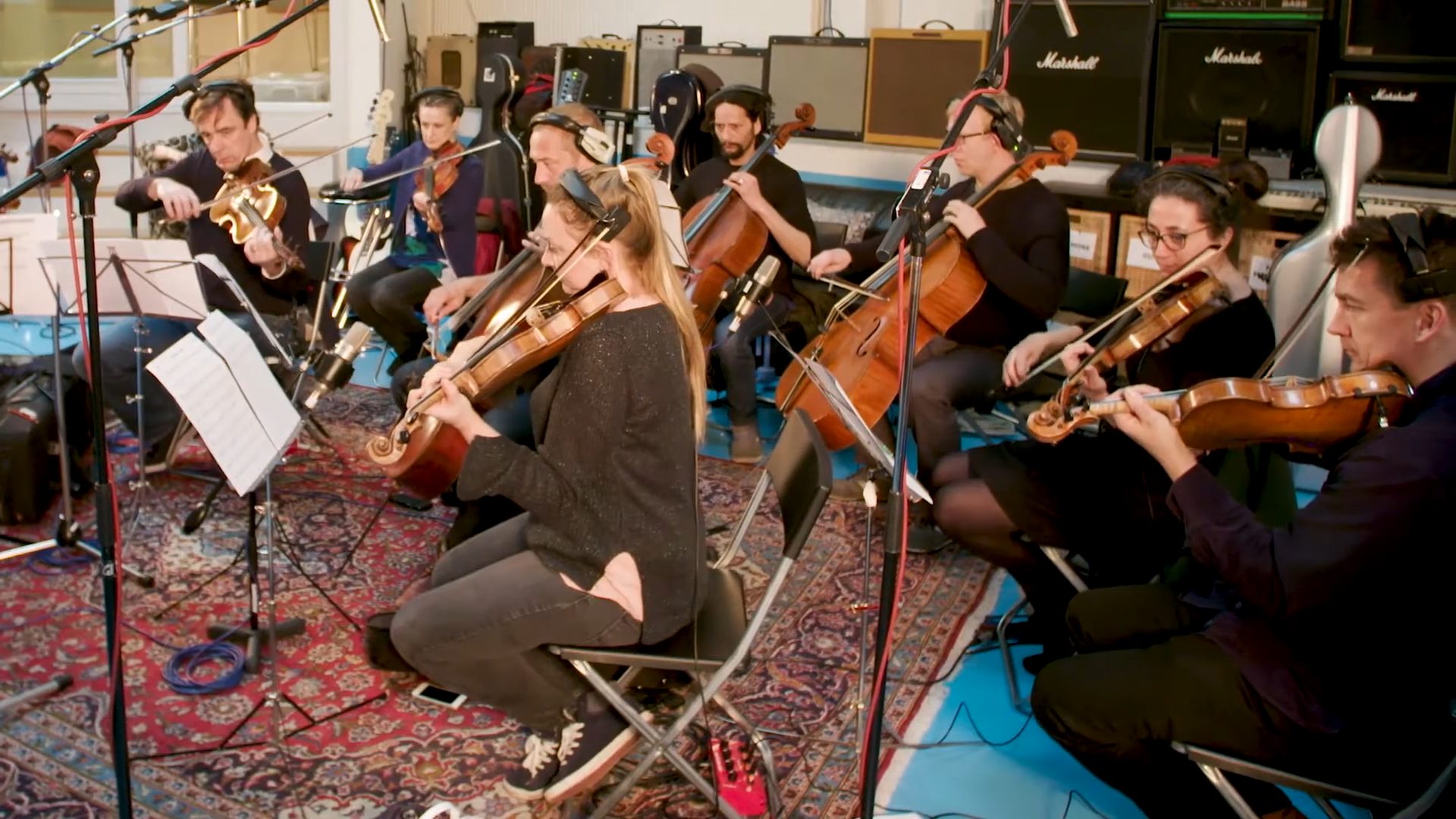 String Orchestra — Studio Recording Session A string orchestra recording in headphones at a studio session, with amplifiers stacked behind.