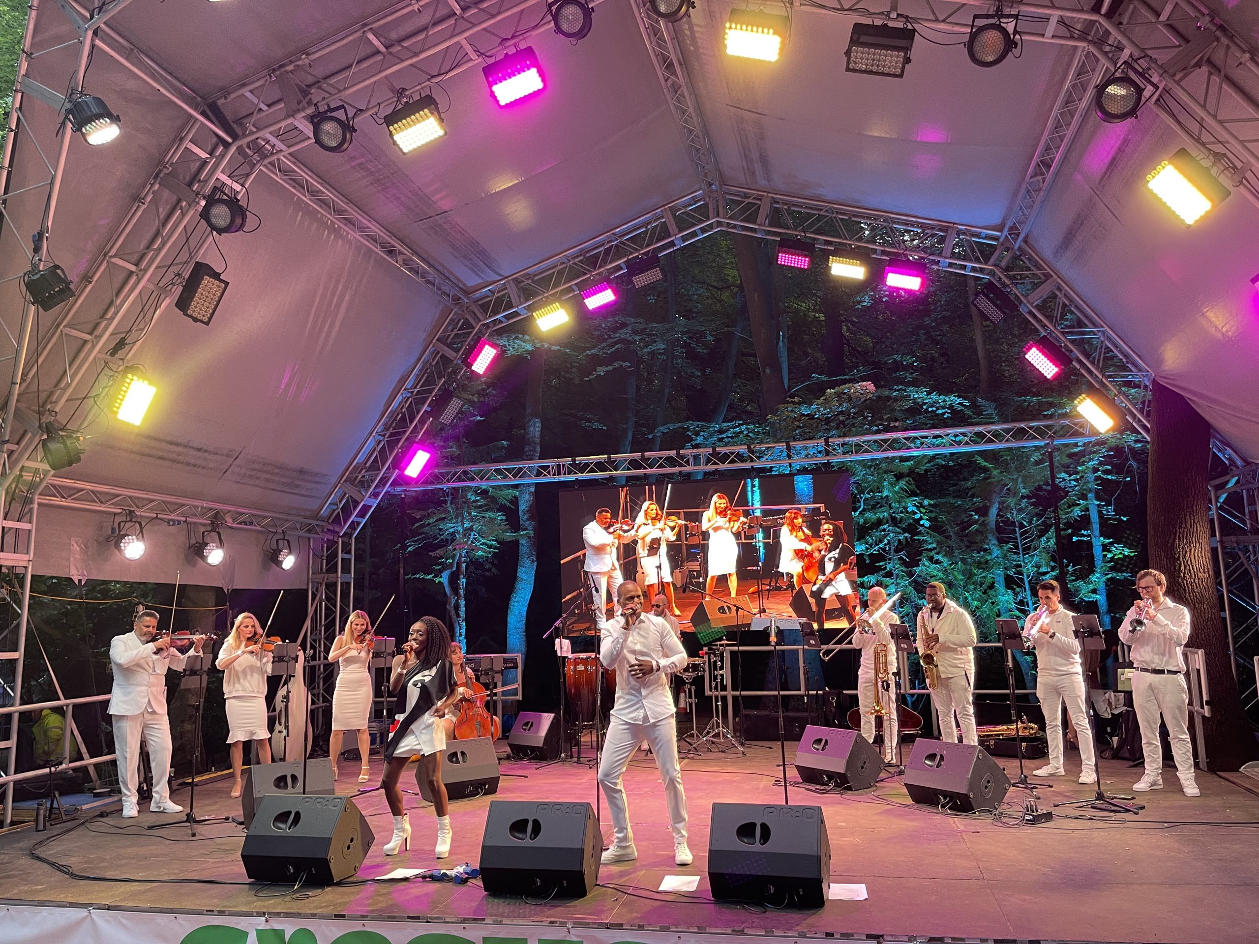 Bangers & Anthems — 13-Piece Live Band on Outdoor Festival Stage Urban Soul Orchestra's Bangers & Anthems band performing on an outdoor stage, strings, horns and vocalists dressed in white.