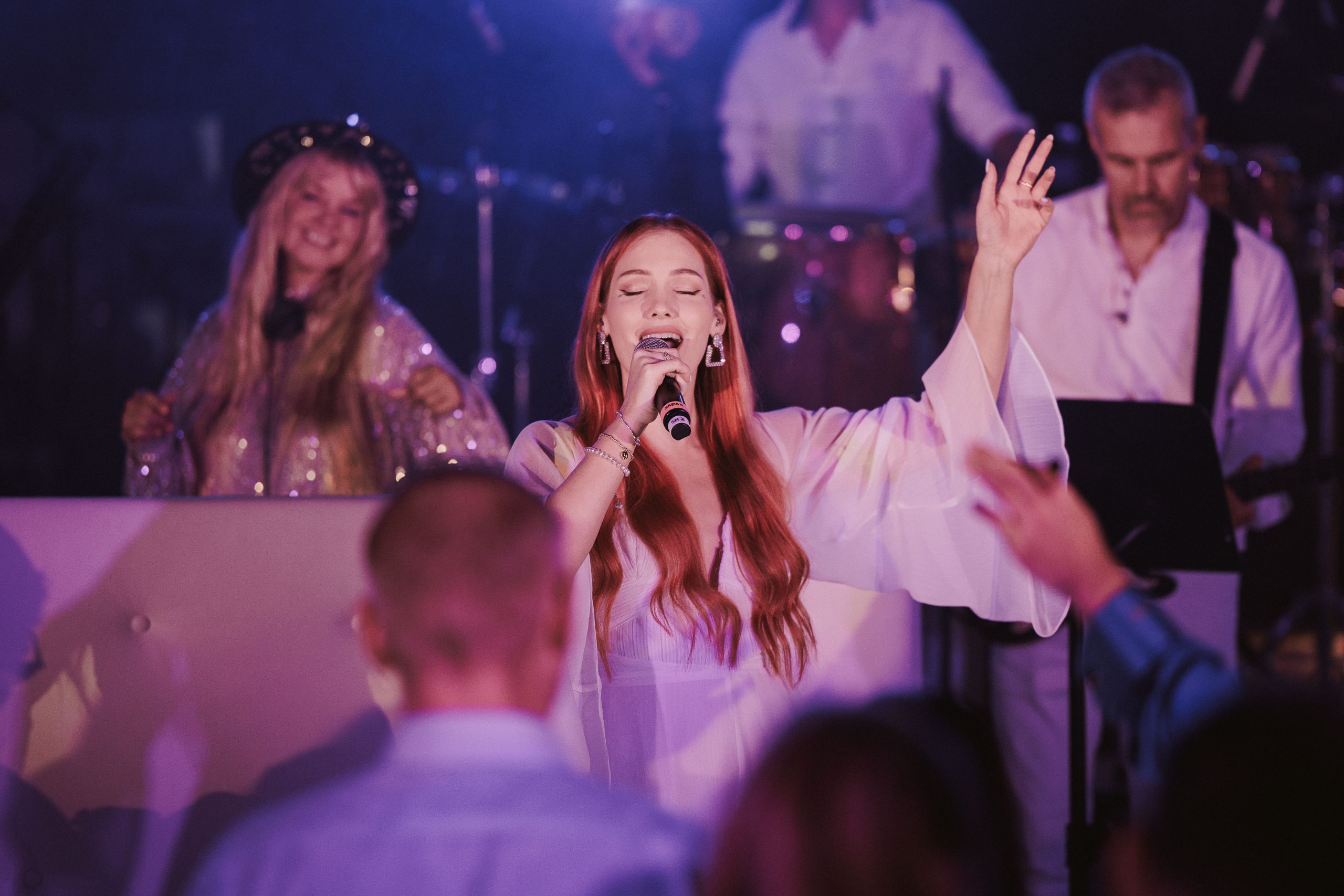 Show Band Vocalist Performing at a Destination Wedding An Urban Soul Orchestra vocalist singing with one arm raised, backed by a DJ, drummer and guitarist under pink and purple stage lighting.