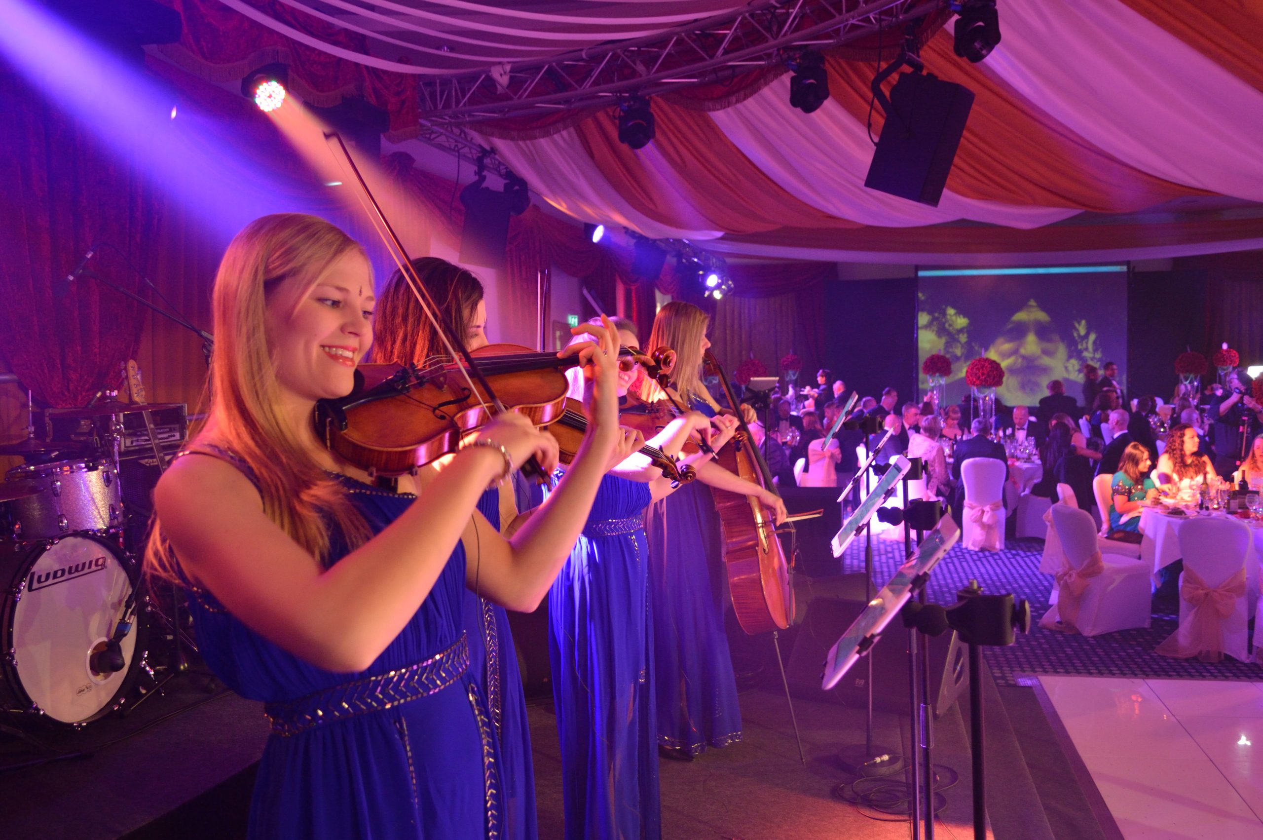 Themed Bands — Bollywood String Quartet at a Charity Ball Four Urban Soul Orchestra string players in blue gowns performing on a lit stage, viewed across floral table arrangements.