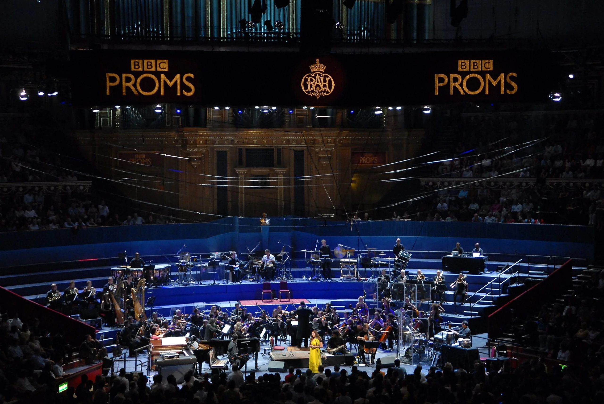 BBC Proms, Royal Albert Hall Orchestra and soloist performing in the arena of the Royal Albert Hall beneath BBC Proms signage, with harps, brass and percussion on the surrounding risers