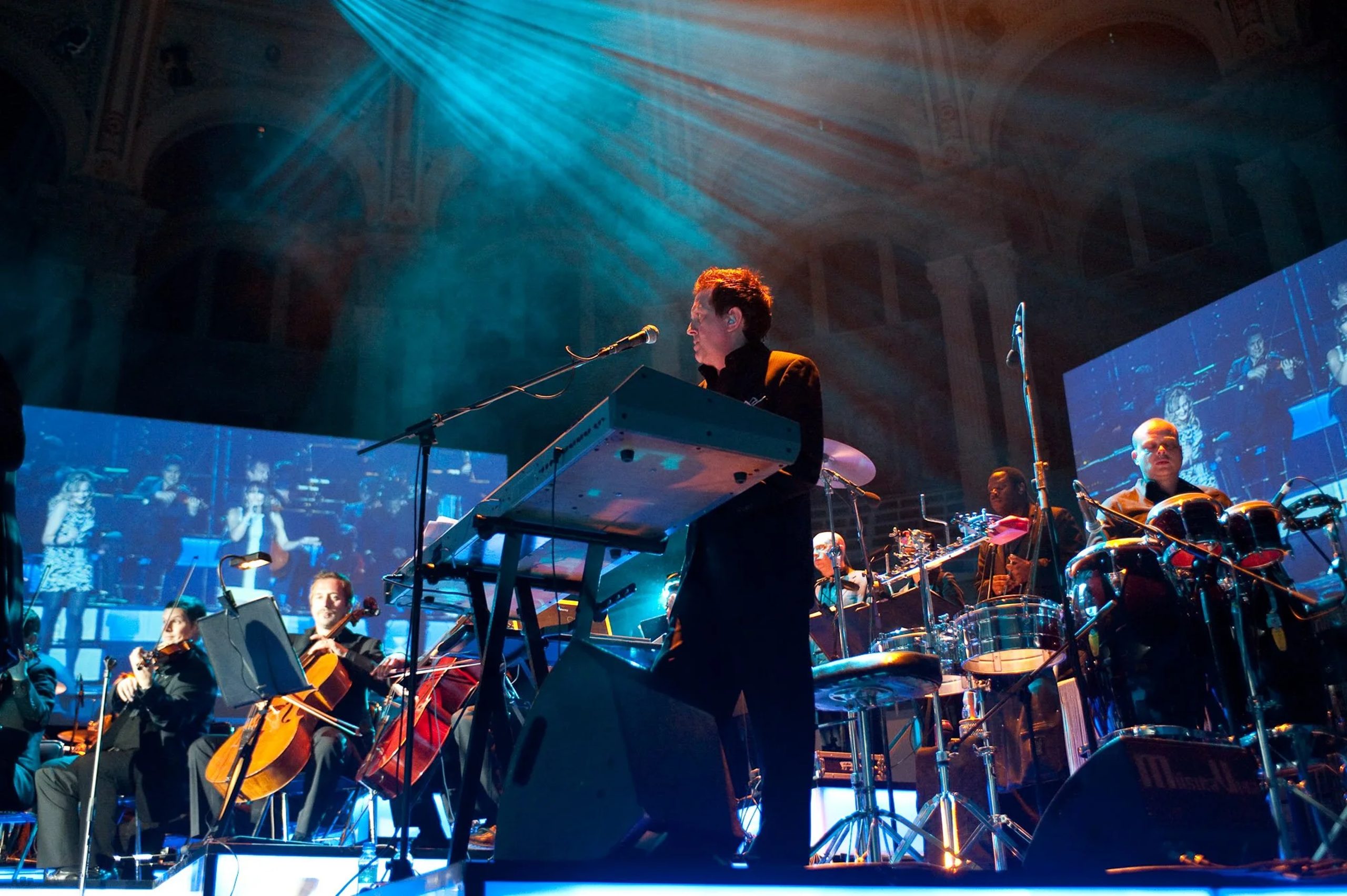 Urban Soul Orchestra performing Musical director at a keyboard with a microphone, leading strings and percussion on stage in a columned hall under blue lighting