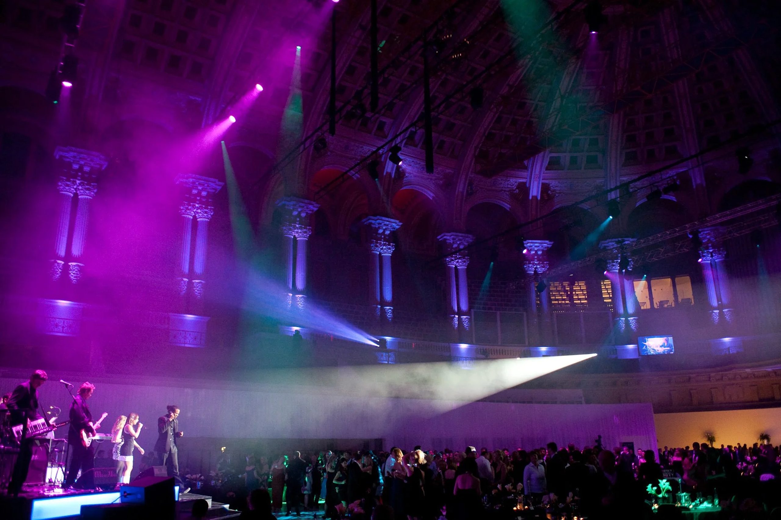 Gala performance Urban Soul Orchestra band performing with vocalists on a stage in a domed hall with Corinthian columns, lit in purple and green, with a seated dinner audience below