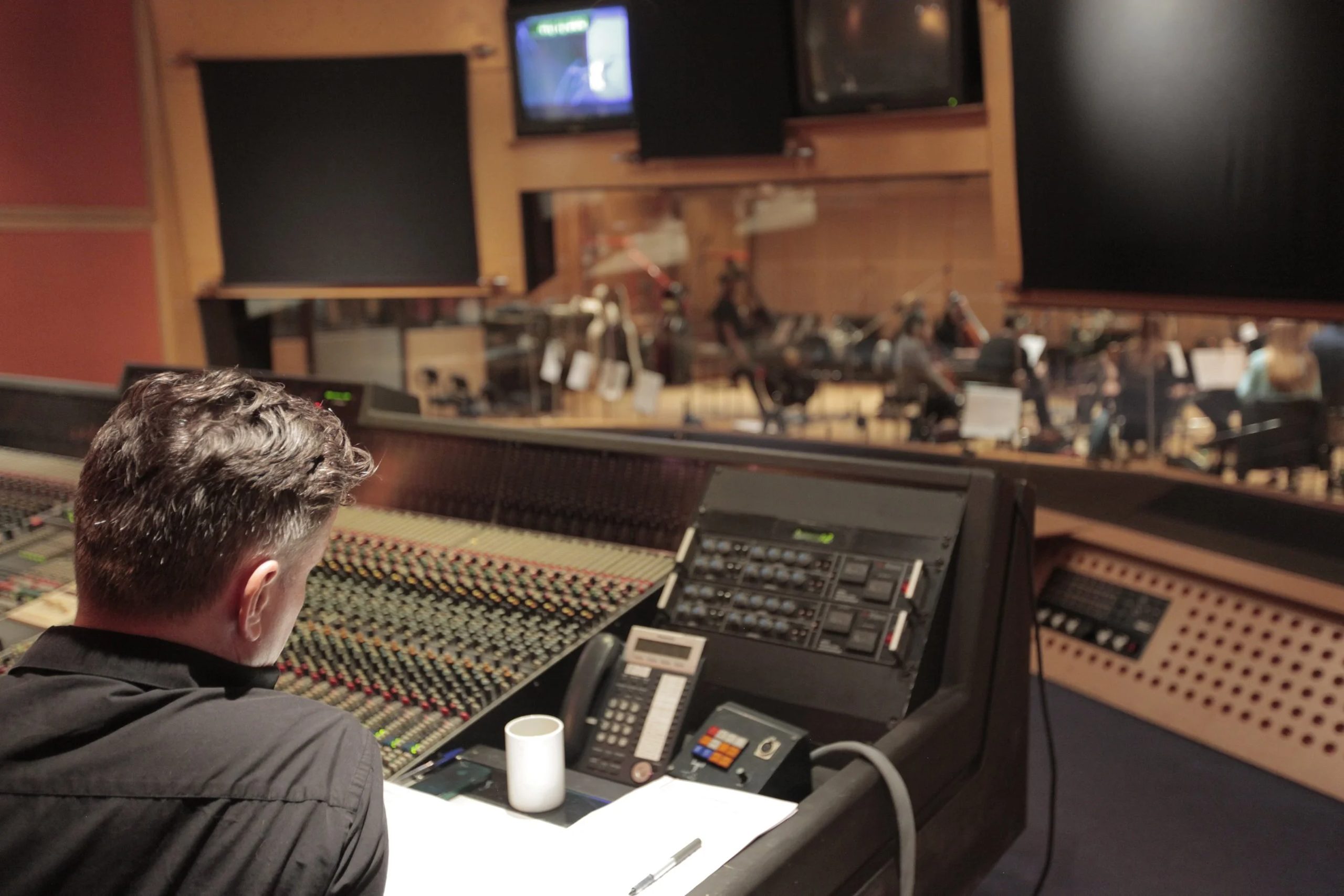 Session recording Recording engineer at a large mixing console in a studio control room, with an orchestra visible through the glass in the live room beyond