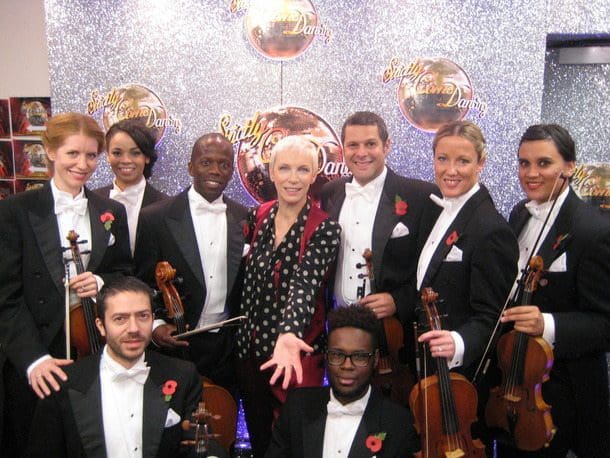 USO String Section with Annie Lennox — Television Performance Eight Urban Soul Orchestra string players in white tie standing with Annie Lennox on a television set.