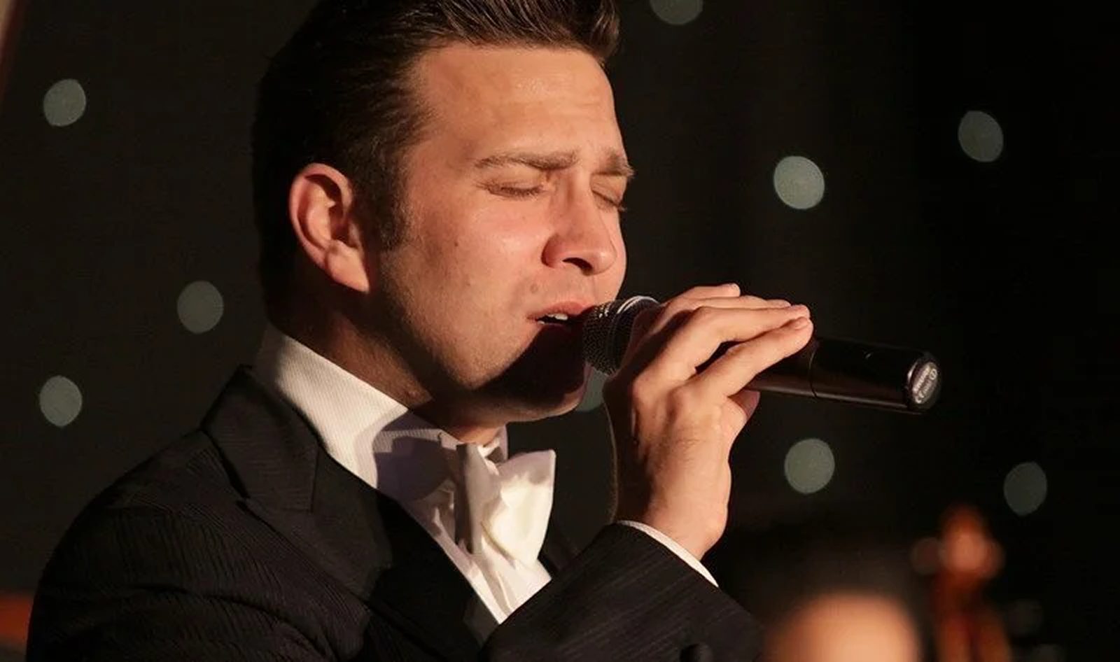 Swing Band vocalist Urban Soul Orchestra Swing Band vocalist in black tie singing into a handheld microphone, eyes closed, in front of a star-cloth backdrop