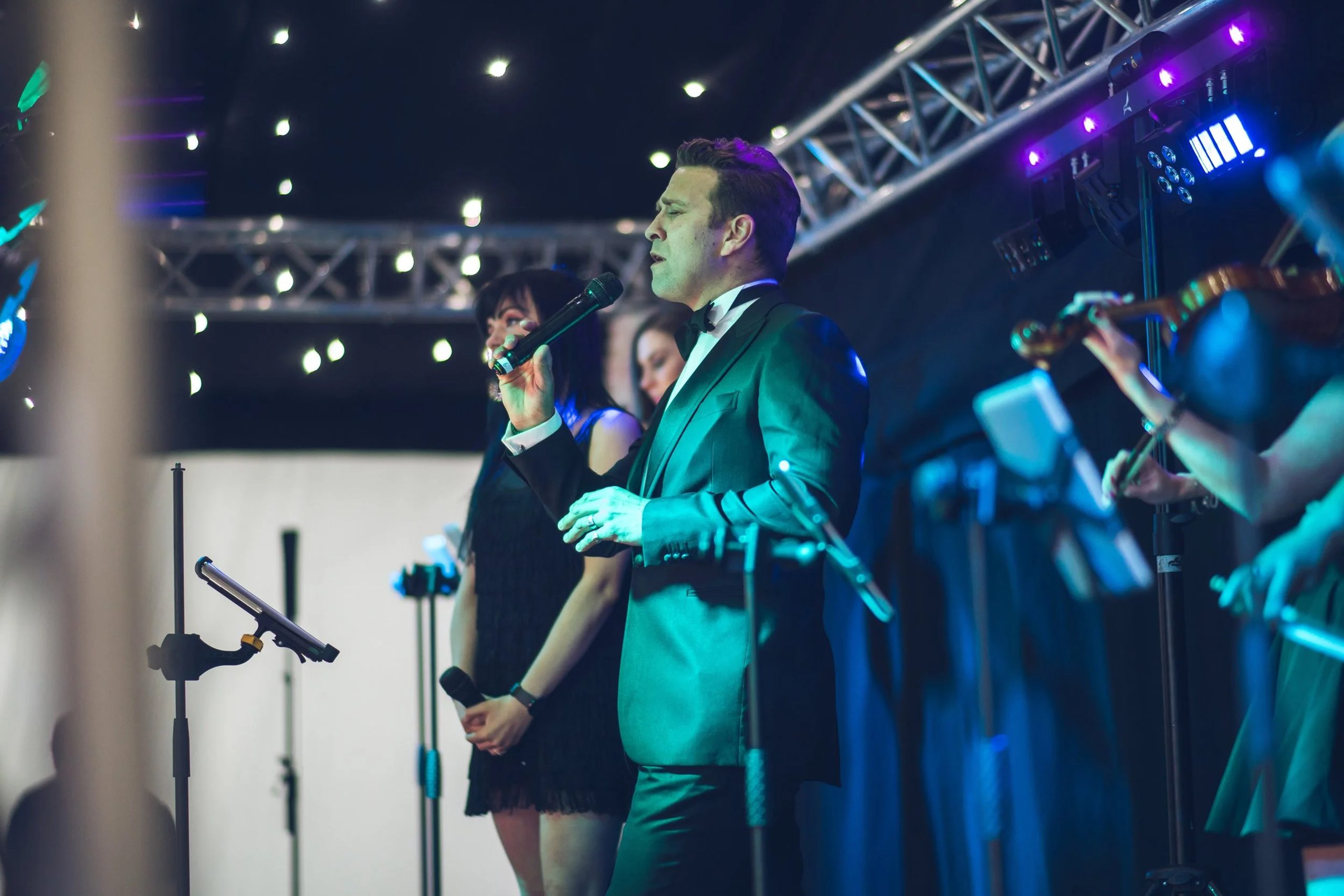 Live band lineup Urban Soul Orchestra vocalist in a teal dinner jacket singing on stage with a backing vocalist and string players under stage lighting
