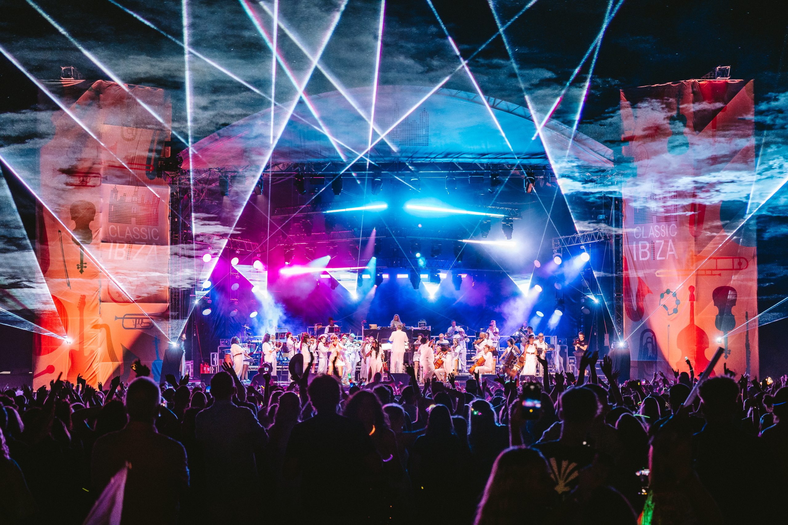 Orchestral Performance at Classic Ibiza, Burghley House 2026 An orchestral ensemble in white performing on a large stage beneath a laser array, with thousands of audience members and smartphones raised.